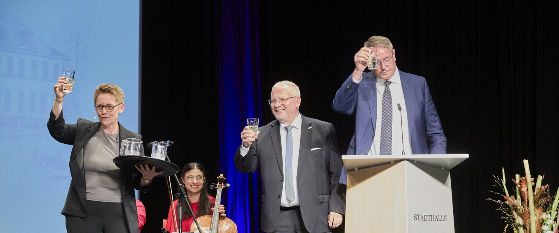 Hans-Ulrich Ihlenfeld (Vorsitzender des Bezirkstags Pfalz) und Alexander Schweitzer (Ministerpräsident Rheinland-Pfalz) stoßen auf der Bühne mit Weinschorle an